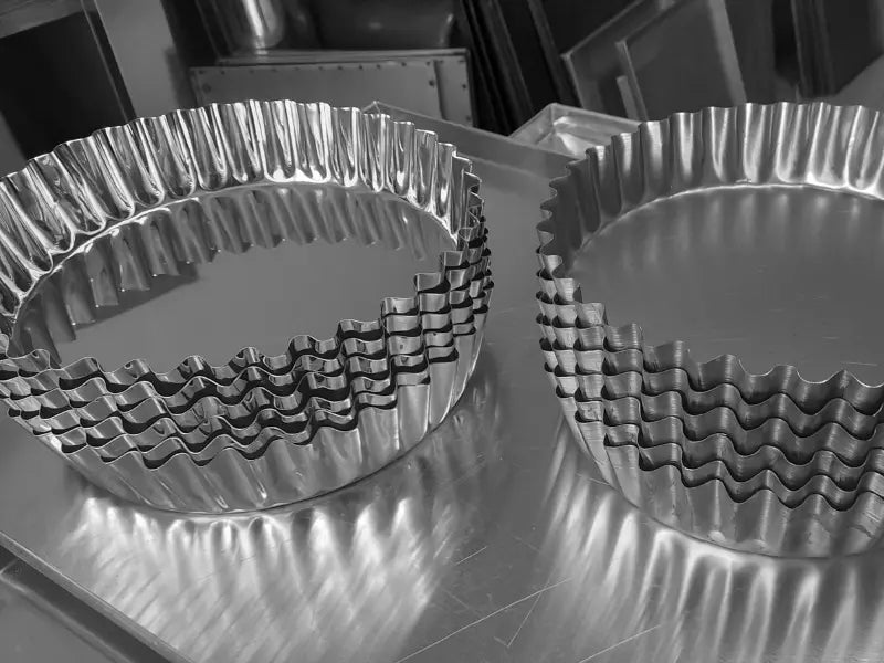 Două stive de inele profesionale din inox pentru tartă și quiche, cu margini canelate (ondulate). Echipamente de înaltă calitate pentru laboratoare de patiserie, așezate pe o masă de lucru din atelier.