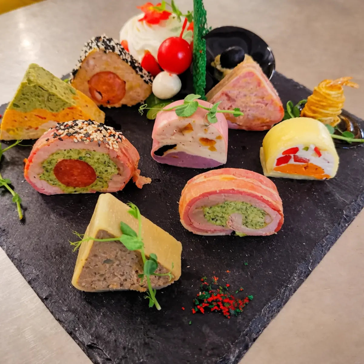 Prezentare de tip finger food cu rulouri și felii de terină cu forme geometrice, așezate elegant pe un platou negru din ardezie.