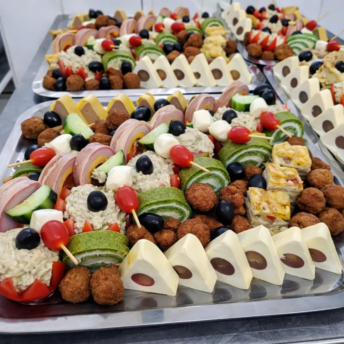 Platouri mari de catering încărcate cu aperitive variate și terine realizate în forme triunghiulare din inox, pregătite pentru evenimente.