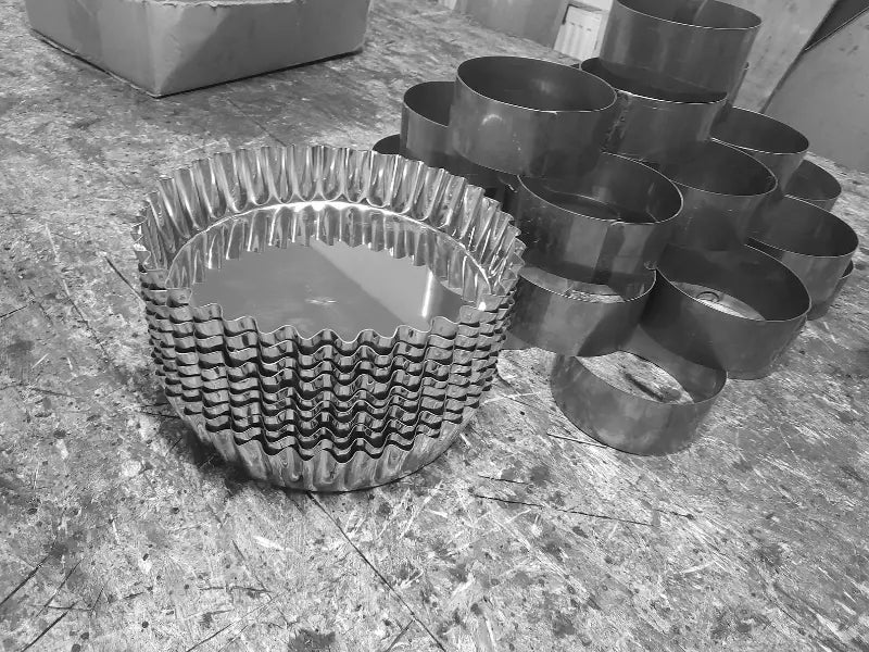 Stivă de forme profesionale tip tavă tartă cu margini canelate din inox, așezate pe o masă de lucru din atelierul de producție, alături de alte inele metalice simple pentru patiserie.
