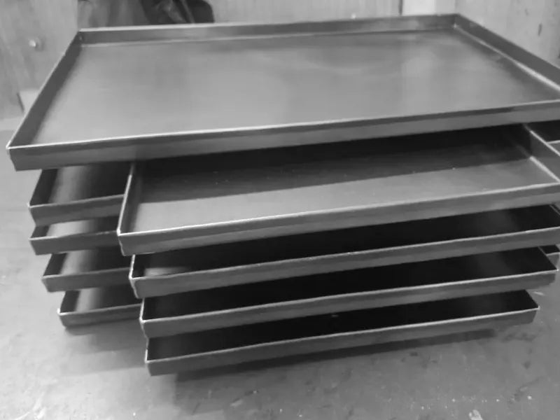 Detaliu cu tăvi de copt profesionale din tablă neagră, suprapuse în atelier. Aceste tăvi robuste de 60x40 cm sunt concepute pentru utilizare zilnică intensă în brutării, patiserii și bucătării comerciale.