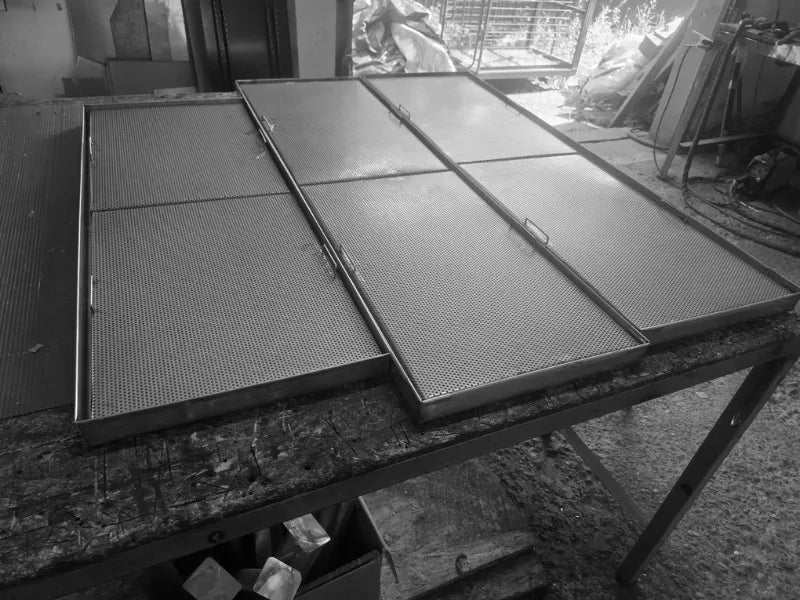 Mai multe tăvi perforate din inox AISI 304 pentru scurgerea paharelor, fotografiate pe o masă de lucru din atelier. Având dimensiunile standard de 600x400 mm, aceste echipamente profesionale robuste sunt gata să echipeze complet stațiile de lucru din baruri și locații HoReCa.